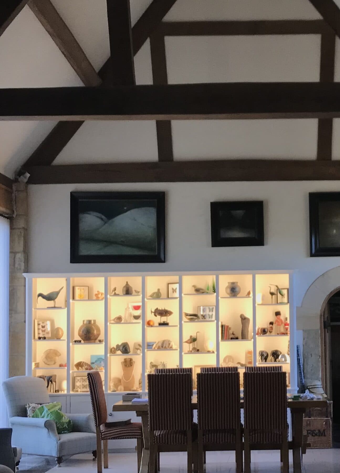 Barn conversion dining room with exposed oak trusses and a wall of lit display shelving beside the dining table