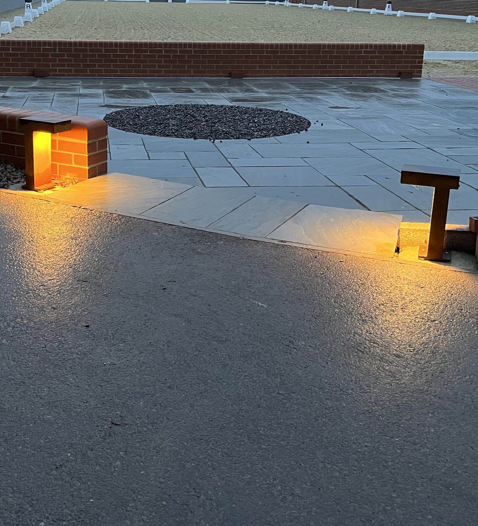Sandstone patio and driveway lit at dusk by recessed lights set into a brick pier and a bollard light beside the steps