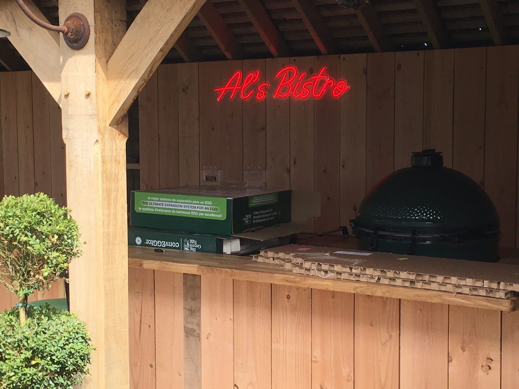 Covered outdoor kitchen in oak with a neon bistro sign lit above the worktop and a wall light on the corner post