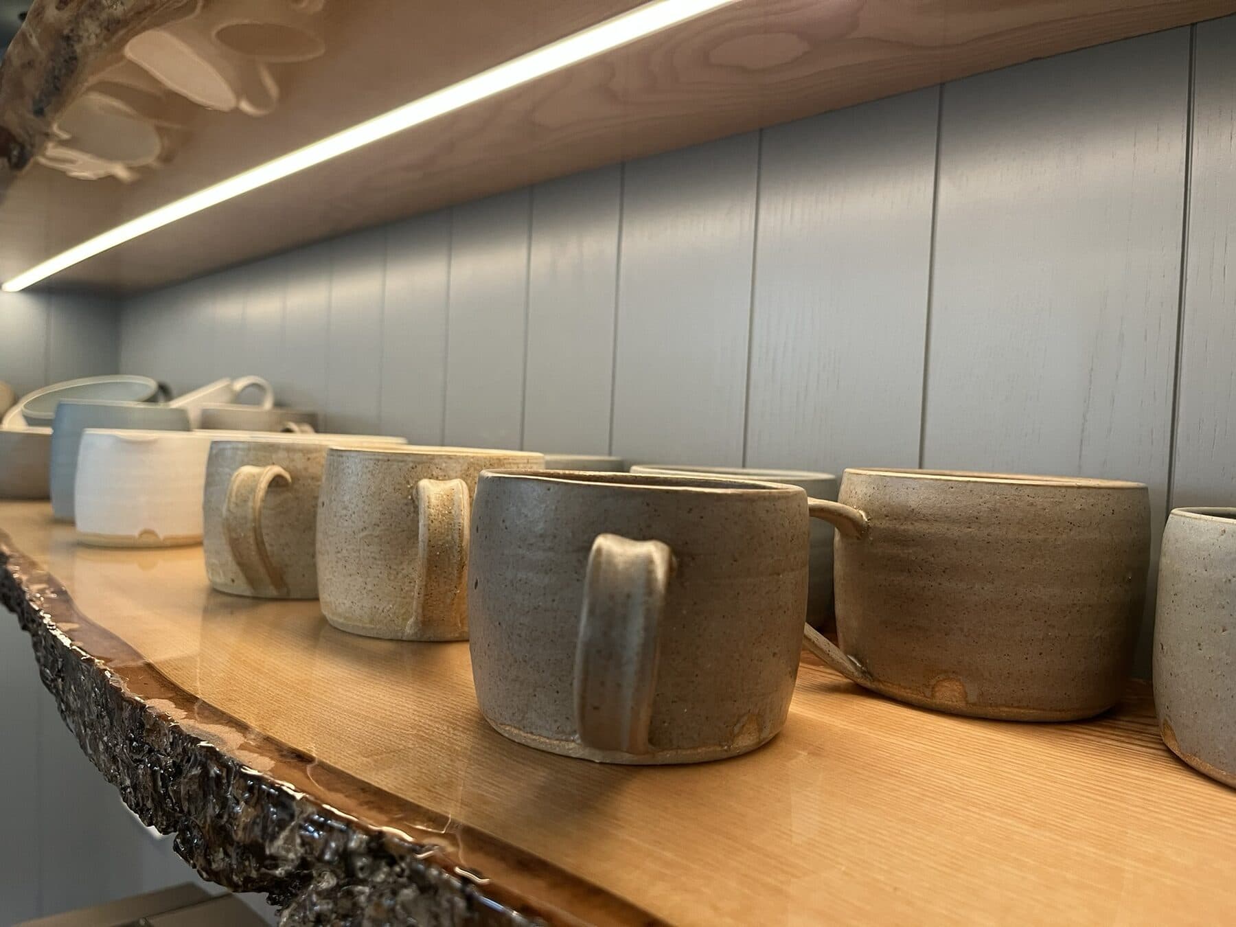 Close up of a warm LED strip lighting a live edge oak shelf holding handmade pottery mugs